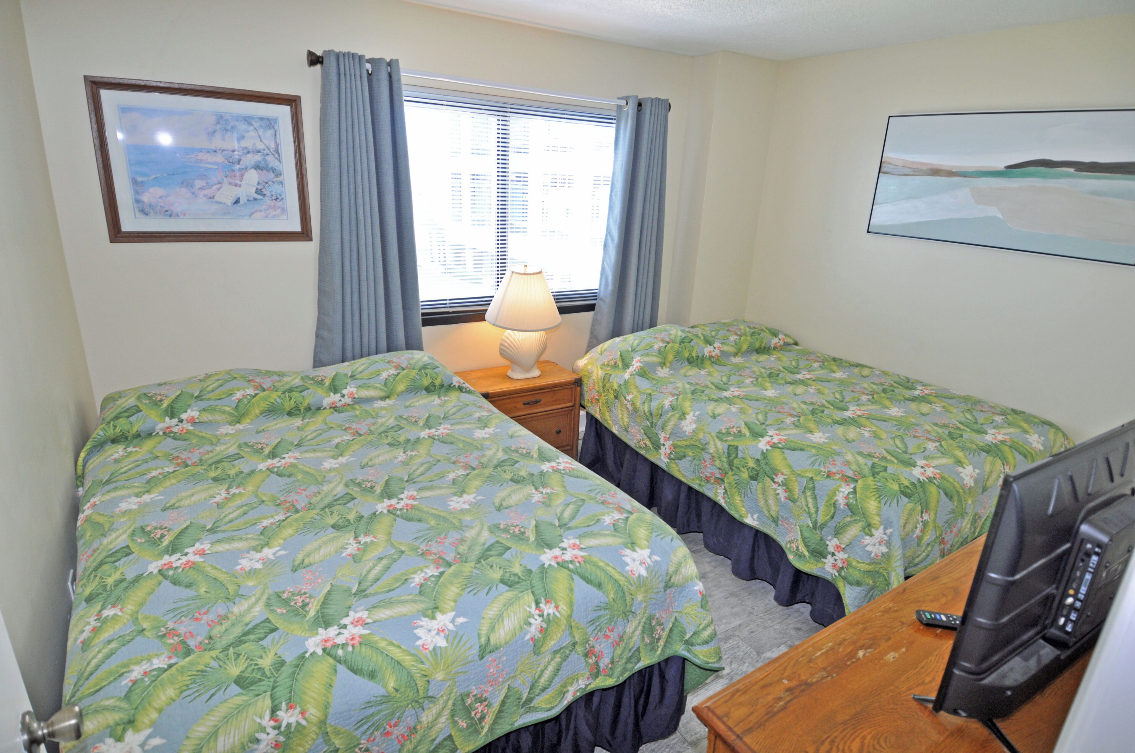 A bedroom with two beds, a nightstand with a lamp, a TV, and a window with curtains.