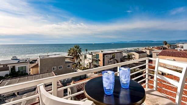 A balcony with a table and two glasses, overlooking a coastal cityscape with buildings and the sea. - Beautiful Views - Sand Section of Manhattan Beach  (Manhattan Beach)