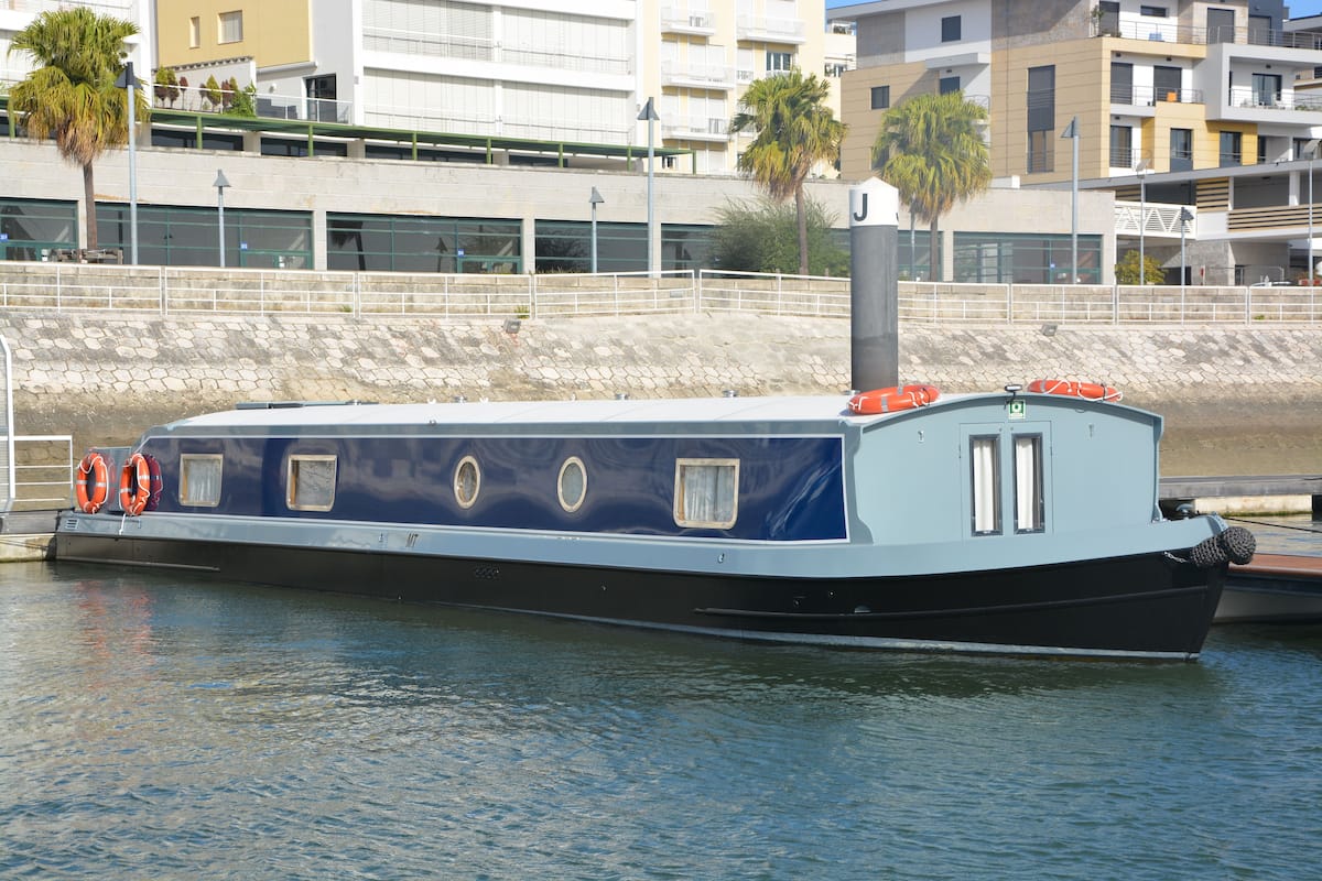 Un barco estrecho amarrado en un puerto deportivo, con un muro de piedra y edificios al fondo.
