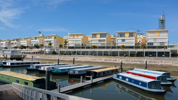 Een jachthaven met verschillende boten afgemeerd, een promenade langs het water met moderne gebouwen, en een heldere lucht.