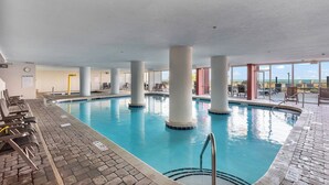 Piscina interna com água azul cristalina, rodeada por pilares brancos, e vista para a praia através de grandes janelas.