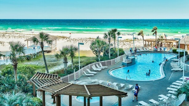 A pool area with a view of the beach, lounge chairs, and a wooden deck.