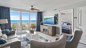 A spacious living room with a large TV, a sofa, a chair, and a balcony with a view of the sea.
