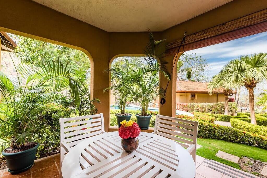 A covered patio with a round table and chairs, surrounded by potted plants and a well-maintained garden.
