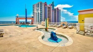 A rooftop pool area with a water fountain, lounge chairs, and a view of the ocean and buildings. - Oceanfront 3BR/3B Lockout. Resort Amenities, Pools, Lazy River, Kiddie Pool (NO. MYRTLE BEACH)