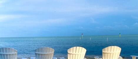 A row of white Adirondack chairs facing a beach with a calm sea and a clear sky.