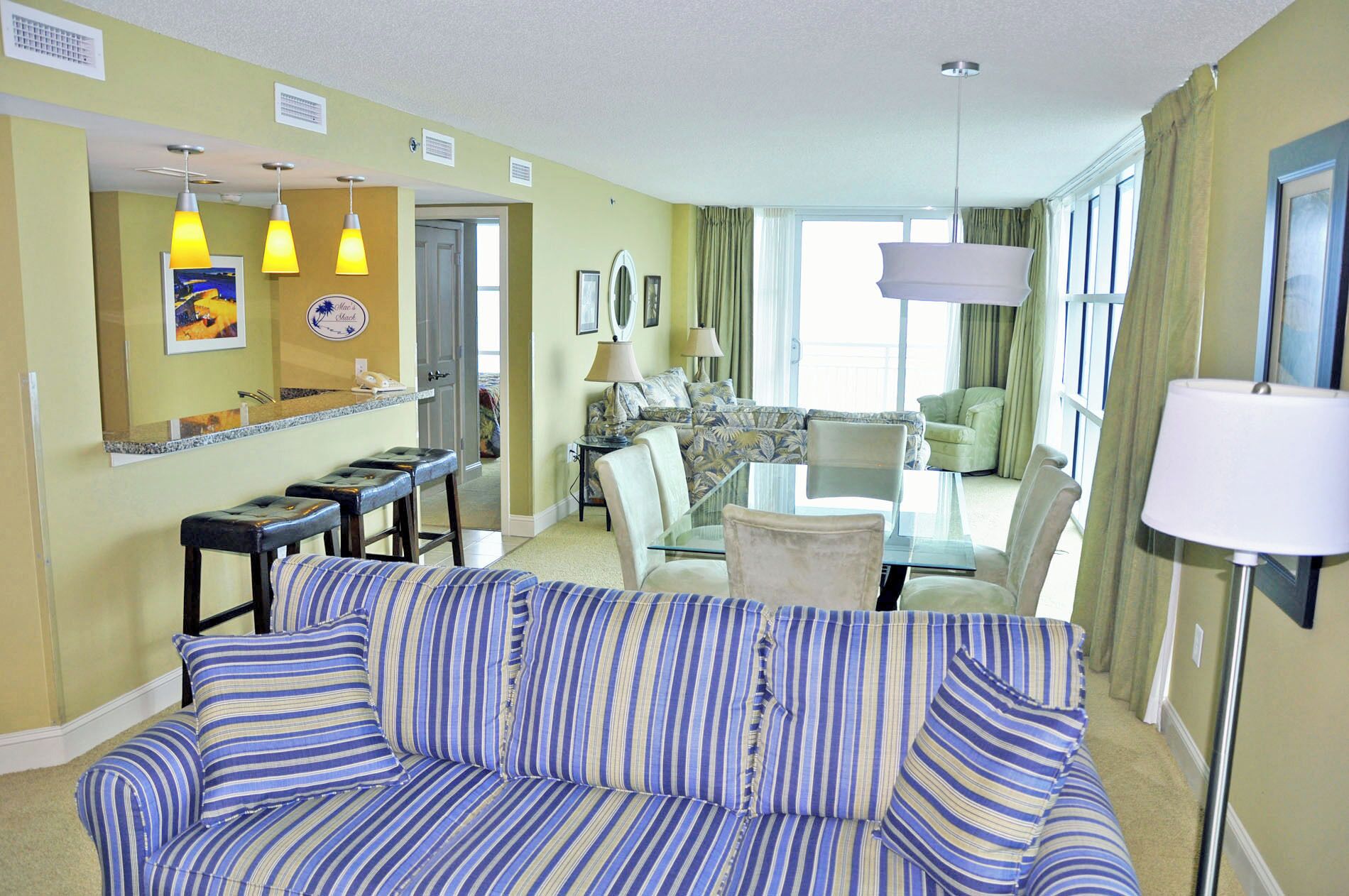 Quarto de hotel com sofá listrado, área de refeições com mesa de vidro e uma janela grande com cortinas.