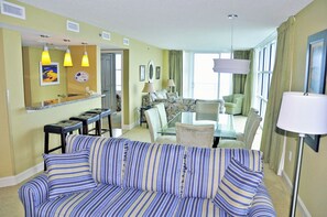 Une chambre d’hôtel avec un canapé à rayures, un coin repas doté d’une table en verre et une grande fenêtre avec des rideaux.