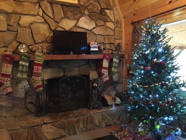 A stone fireplace with a flat-screen TV above it, a Christmas tree with lights and ornaments, and stockings hanging on the mantel.