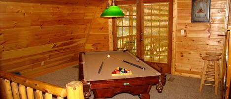 A pool table in a wooden room with a billiards club sign, a green hanging lamp, and a window with blinds.