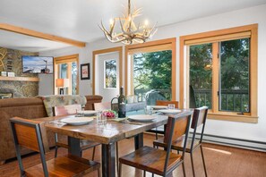 A dining area with a wooden table, chairs, and a chandelier. There are large windows with a view of trees.