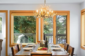 A dining area with a wooden table set for four, a chandelier, and large windows with a view of trees.