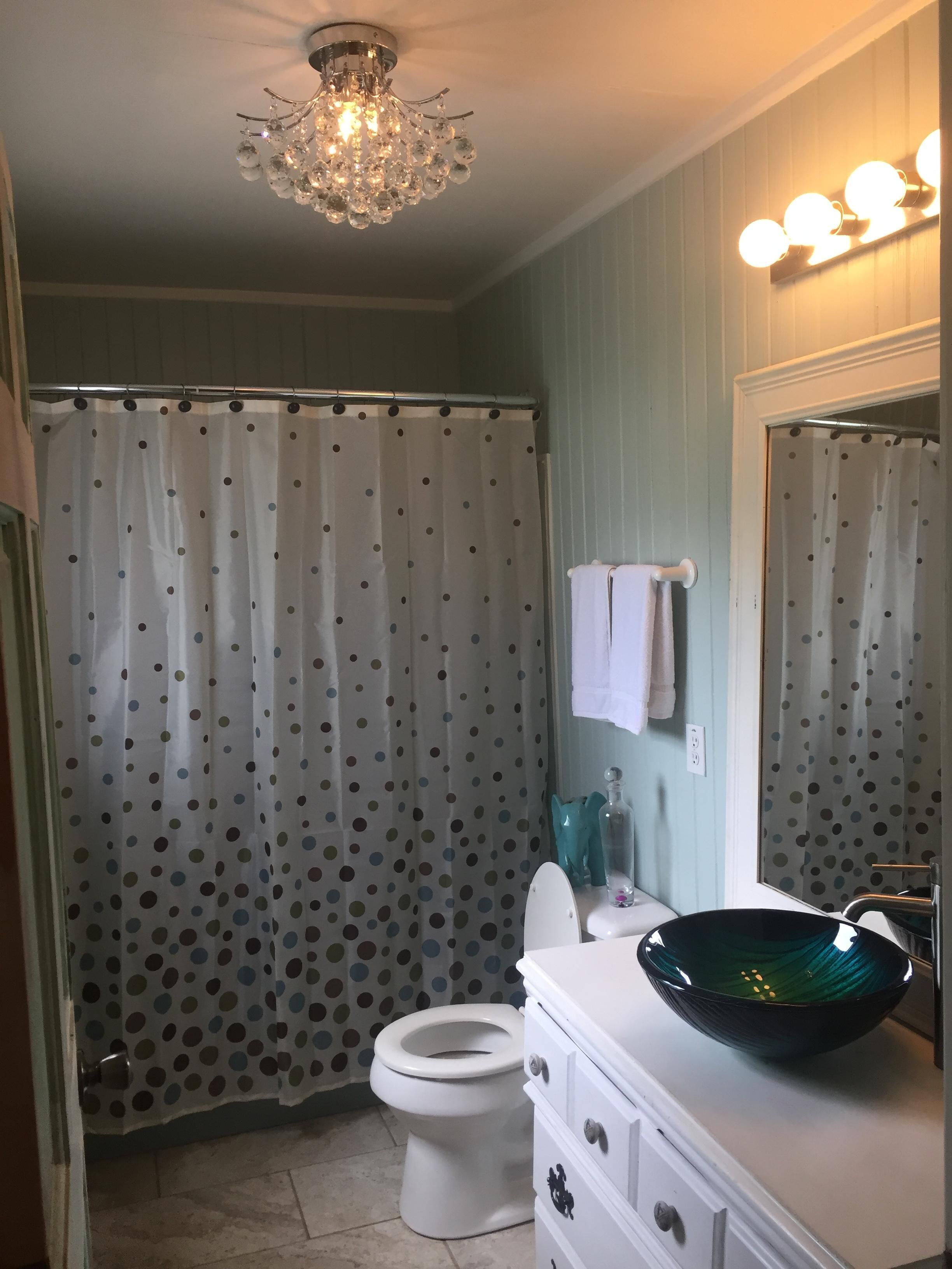 A bathroom with a white toilet, a green glass bowl sink, a chandelier, and a polka dot shower curtain.