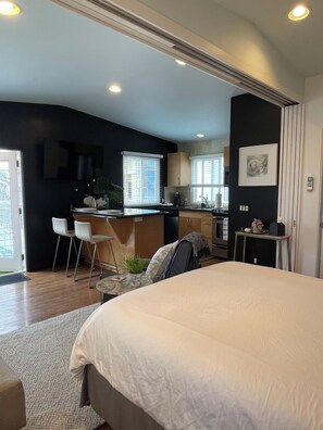 Un dormitorio moderno con una cama grande, un escritorio pequeño y una kitchenette al fondo.
