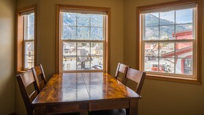 Comedor con mesa y sillas de madera, ventanas amplias con vista a la montaña y una vista de un pueblo con edificios y una estructura roja.