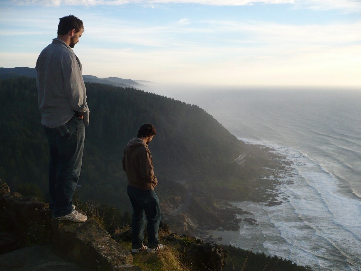 Two people standing on a cliff overlooking a coastal landscape.