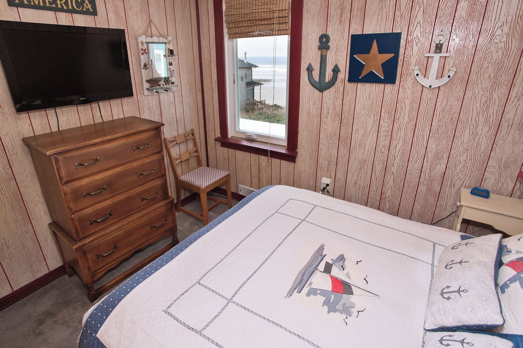 Une chambre avec une commode en bois, une chaise, un lit recouvert d’une courtepointe à motifs marins et une fenêtre donnant sur l’océan.