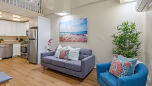 En moderne lejlighed med tekøkken, grå sofa, blå lænestol og stort vindue med udsigt til stranden.