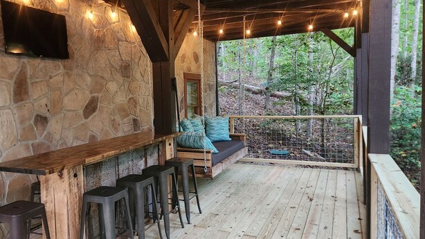 A stone-walled outdoor seating area with a wooden table and bar stools, a swing seat with cushions, and a view of the forest.