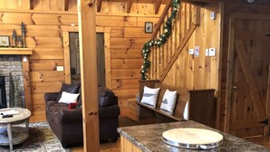 A cozy cabin interior with a wooden interior, a fireplace, a dining area with a round table, and a staircase with a garland.