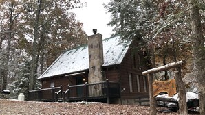 A rustic cabin with a stone chimney, surrounded by trees and covered in snow.