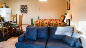 A living room with a blue sofa, a dining area with wooden chairs and a table, and a bed with patterned bedding.