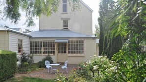 Een gebouw met twee verdiepingen, voorzien van een overdekt terras, een kleine tuin met witte stoelen en een haag.