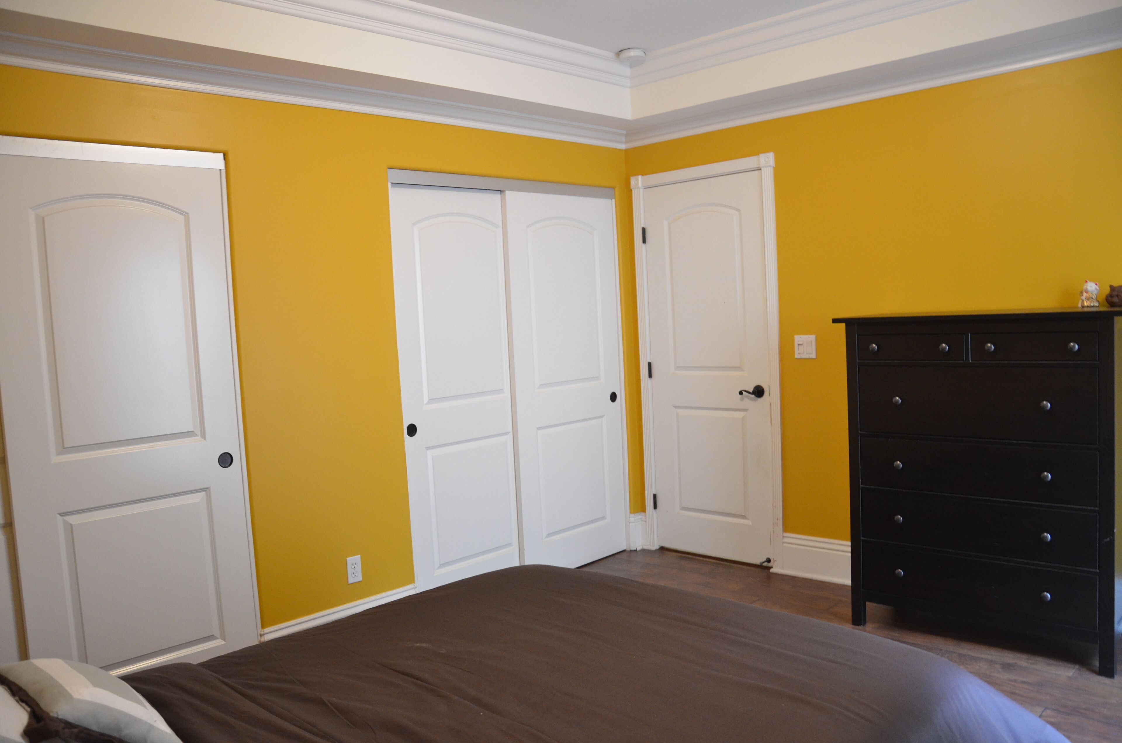 Een slaapkamer met een bed, twee witte deuren, een dressoir en gele wanden.