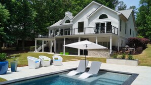 A pool area with lounge chairs, a covered patio, and a large house in the background. - 5 BDRM Beautiful Peaceful Lake Front Home 3 Kings, 1 Queen, 4 Twins, Pool, Spa (Greensboro)