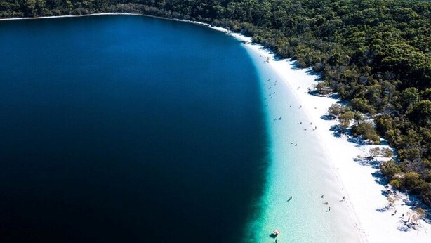 Vista aérea de una playa con mar azul cristalino, arena blanca y colinas boscosas al fondo.