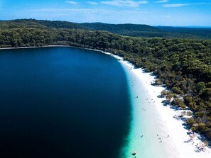 An aerial view of a beach with a clear blue sea, white sand, and dense forested hills in the background.