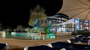Zona de piscina con sillas de descanso, un espacio de estar techado y un edificio moderno con techo curvo.
