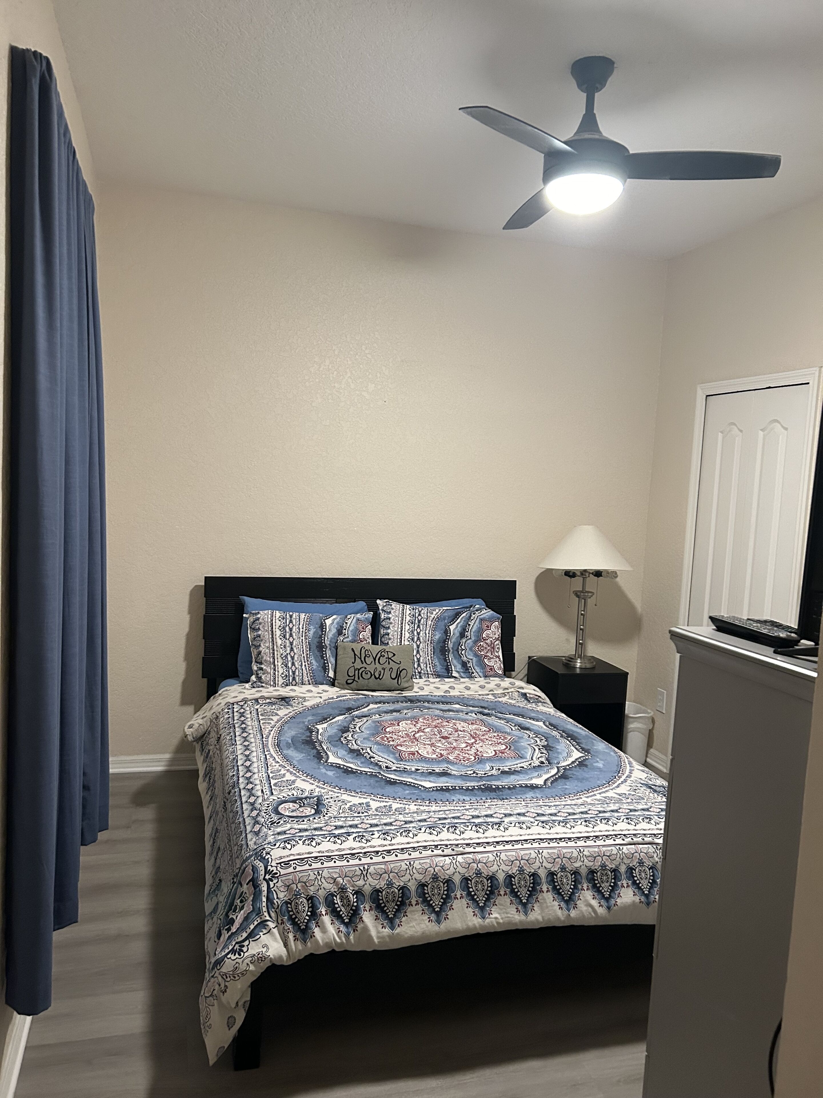 Ein Schlafzimmer mit Bett, Deckenventilator, Nachttisch, Lampe und einer Tür.