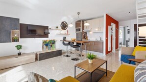 Ein modernes Wohnzimmer mit Sofa, Couchtisch und an der Wand montiertem Fernseher. Es gibt eine Küchenzeile mit Mikrowelle, Backofen und Spüle.