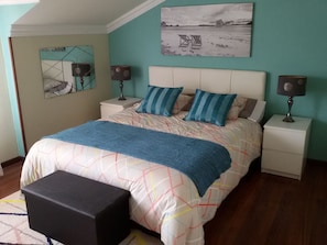 A bedroom with a bed, two bedside tables, a nightstand, a black ottoman, a wall-mounted mirror, and a beach-themed painting.