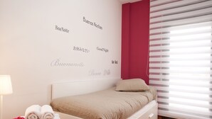 A bedroom with a bed, a bedside table with a lamp and towels, a wall with multilingual "Good Night" messages, and a window with blinds. - SUNNY ATTIC, 2 TERRACES, PRIVATE SOLARIUM, 3 ROOM, 2 BATHS. 3 MIN TO SUBWAY. (Barcelona)