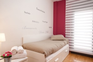 A bedroom with a bed, a bedside table with a lamp and towels, a wall with multilingual "Good Night" messages, and a window with blinds.