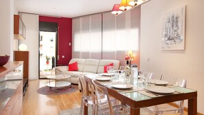 A modern living room with a dining area, featuring a glass dining table, clear chairs, and a sofa with red cushions. - SUNNY ATTIC, 2 TERRACES, PRIVATE SOLARIUM, 3 ROOM, 2 BATHS. 3 MIN TO SUBWAY. (Barcelona)