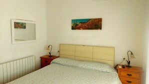 Un dormitorio con cama, mesita de noche, cómoda, aire acondicionado instalado en la pared, espejo y un cuadro.