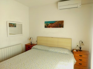 Quarto com cama, mesa de cabeceira, cômoda, ar-condicionado fixado na parede, espelho e pintura.