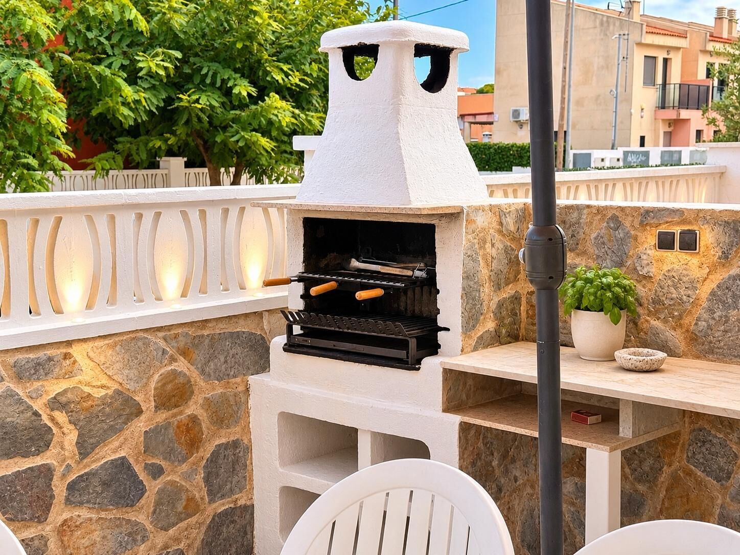 Una parrilla para barbacoa al aire libre con un muro de piedra de fondo, una silla blanca y una planta en maceta.