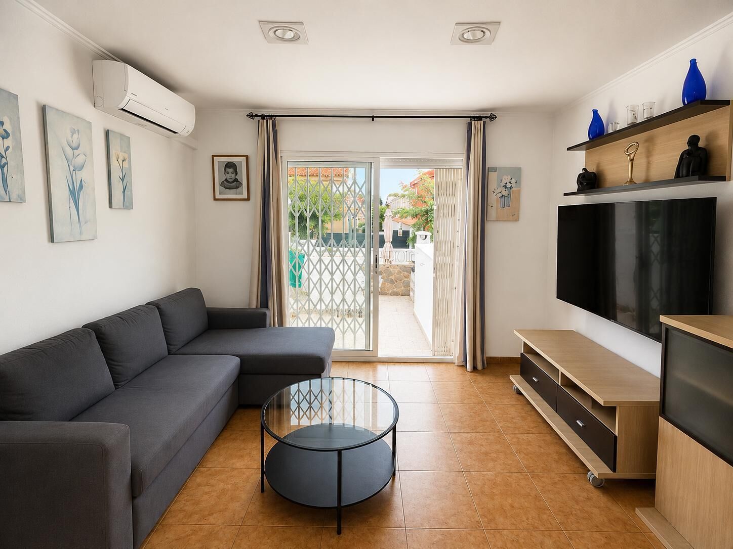 A modern living room with a grey sofa, a glass coffee table, a flat-screen TV, and a wooden TV stand.