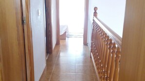 Un pasillo con barandal de madera, suelo de baldosas y una puerta que da a una habitación con cama.