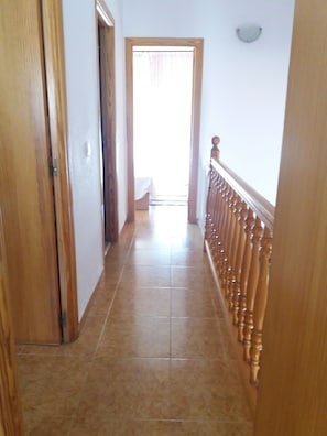 Um corredor com corrimão de madeira, piso ladrilhado e uma porta que dá para um quarto com cama.