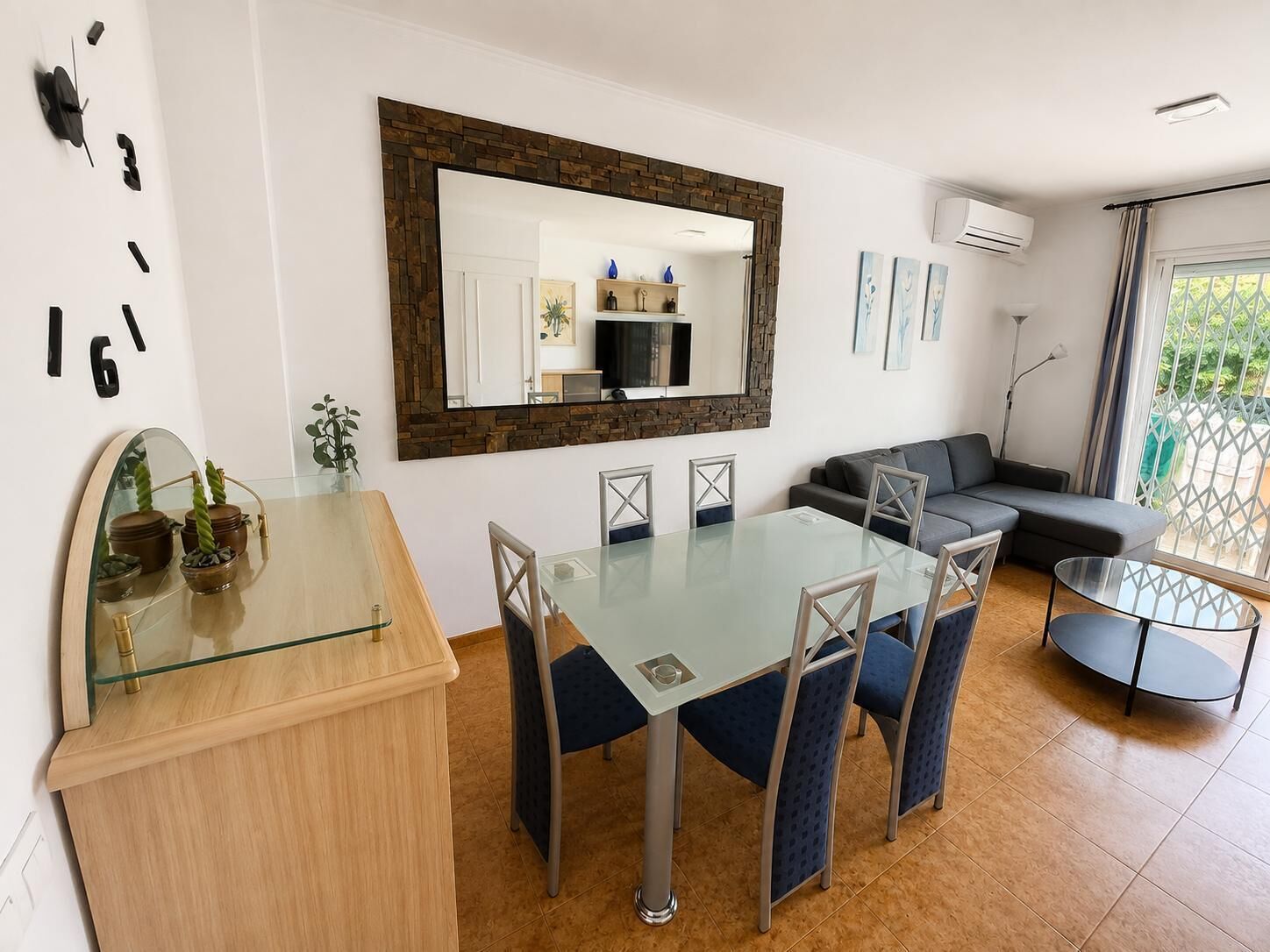 A modern living room with a glass dining table, a sofa, and a large mirror.