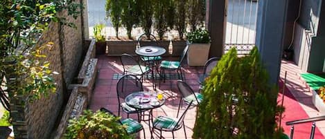 Un patio al aire libre con mesas y sillas, rodeado de plantas en macetas y un suelo de baldosas rojas.