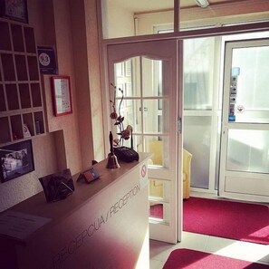 A reception desk with a sign reading 'RECEPTION,' a framed certificate on the wall, and a door leading to a room with a red carpet. - Guest Accommodation Majesty (Niš)