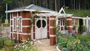 Ein rot backengefliestes Gebäude mit einer weißen Tür und Fenstern, umgeben von einem Garten mit verschiedenen Blumen und einem weißen Zaun.