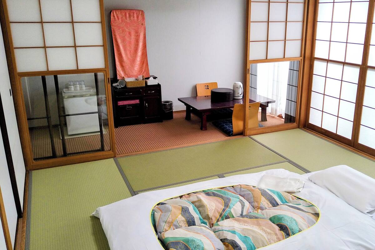 Ein traditioneller japanischer Raum mit Tatami-Bodenbelag, einem Bett mit bunter Decke, einem Schreibtisch mit Wasserkocher und Schiebetüren.