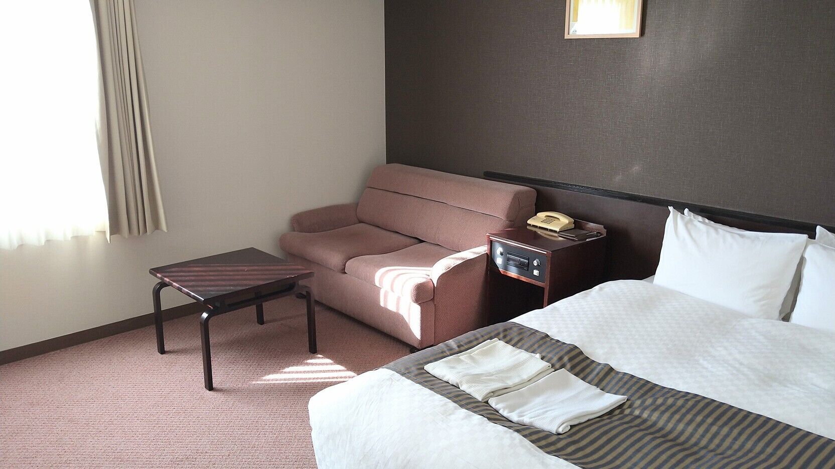 A hotel room with a bed, a sofa, a small table, a nightstand with a phone, and a wall-mounted light.